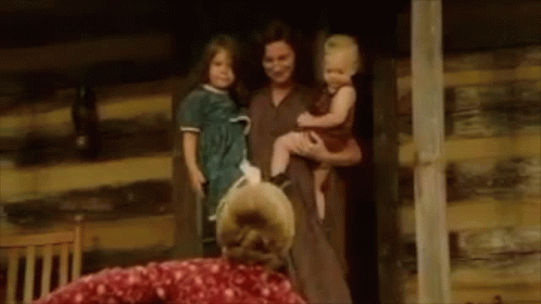 A Woman Is Holding A Baby And A Dog In Her Arms While Standing Next To A Log Cabin . GIF