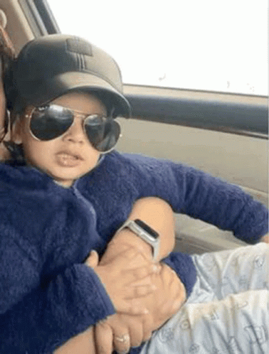 A Woman Is Holding A Baby In Her Arms In A Car . The Baby Is Wearing Sunglasses And A Baseball Cap . GIF