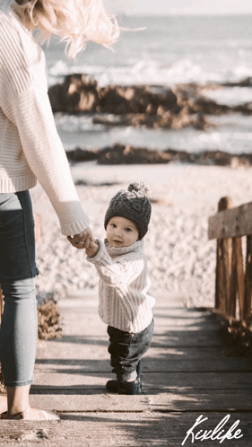 A Woman Is Holding A Baby 'S Hand While They Walk On A Boardwalk GIF