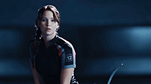 A Woman Is Holding A Bow And Arrow With The Word Hunger Games On Her Shirt GIF