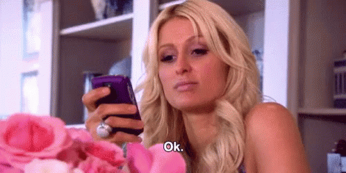 A Woman Is Holding A Cell Phone In Her Hand While Sitting In Front Of A Bouquet Of Pink Roses . GIF