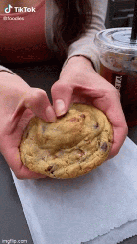 A Woman Is Holding A Cookie In Her Hands With A Cup Of Coffee In The Background . GIF