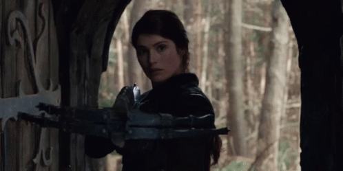A Woman Is Holding A Crossbow In Front Of A Door In A Forest . GIF