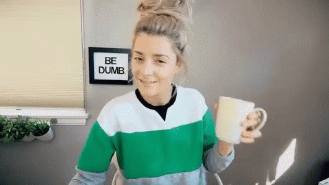 A Woman Is Holding A Cup In Front Of A Sign That Says Be Dumb . GIF