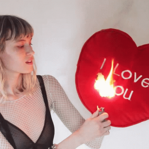 A Woman Is Holding A Lighter Next To A Red Heart Shaped Pillow That Says I Love You GIF