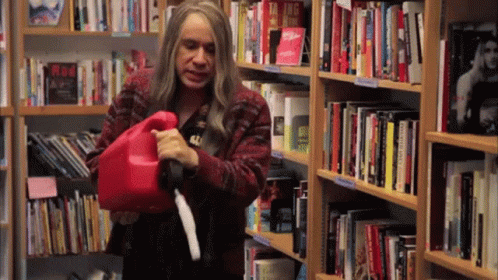 A Woman Is Holding A Red Gas Can In Front Of A Bookshelf With A Book Titled Taking Risks On It GIF