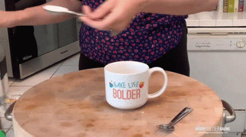 A Woman Is Holding A Spoon Over A Mug That Says Make Life Bolder GIF