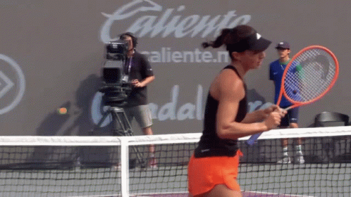 A Woman Is Holding A Tennis Racquet On A Court In Front Of A Wall That Says Caliente GIF