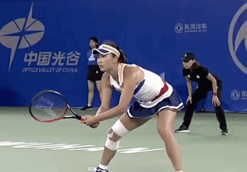 A Woman Is Holding A Tennis Racquet On A Court In Front Of An Optics Valley Of China Banner GIF