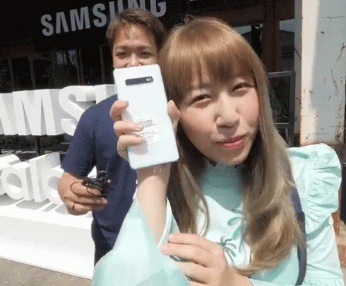 A Woman Is Holding A White Phone In Front Of A Samsung Sign GIF