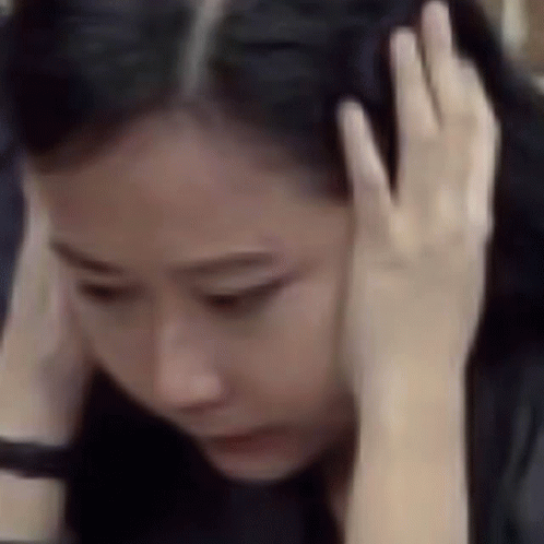 A Woman Is Holding Her Head With Her Hands While Looking Down . GIF