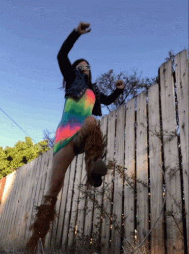A Woman Is Jumping Over A Wooden Fence Wearing A Colorful Dress GIF