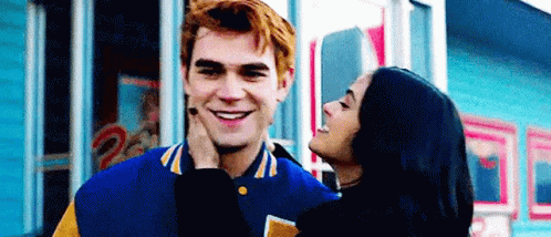 A Woman Is Kissing A Man On The Cheek While They Are Standing In Front Of A Building . GIF