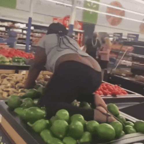 A Woman Is Kneeling Down In Front Of A Bunch Of Green Peppers In A Grocery Store GIF