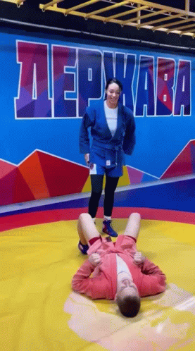 A Woman Is Kneeling On Top Of A Man On A Wrestling Mat In A Gym . GIF