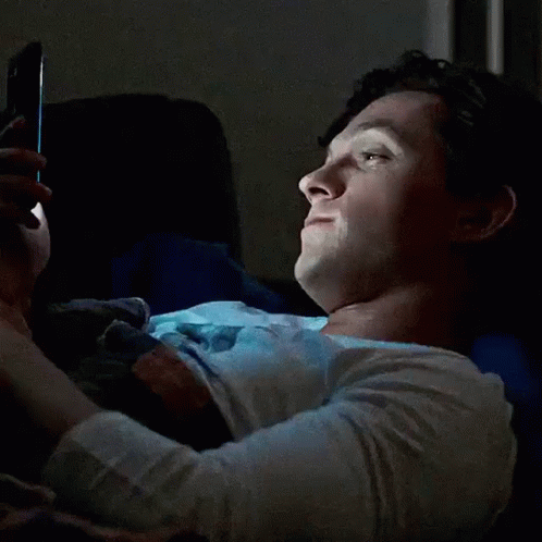 A Woman Is Laying In Bed Looking At Her Phone And Smiling . GIF