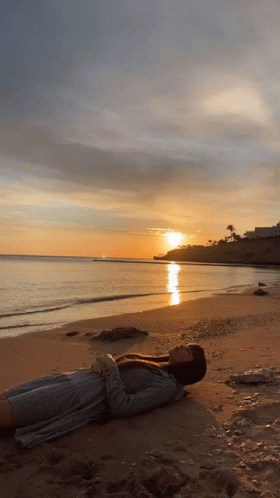 A Woman Is Laying On A Beach With The Sun Setting In The Background GIF