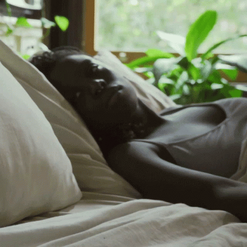 A Woman Is Laying On A Bed With White Sheets And Pillows GIF