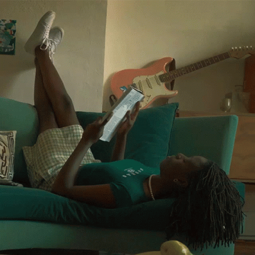 A Woman Is Laying On A Couch Reading A Book With A Guitar In The Background GIF