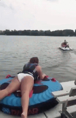 A Woman Is Laying On A Tube In The Water With A Jet Ski Behind Her . GIF