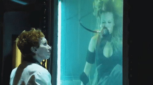 A Woman Is Looking At A Woman In A Tank With The Letter B On Her Chest GIF