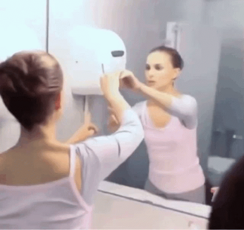 A Woman Is Looking At Her Reflection In A Bathroom Mirror . GIF