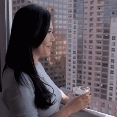 A Woman Is Looking Out A Window With A Cup Of Coffee In Her Hand GIF