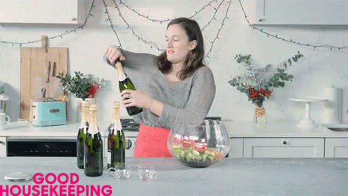 A Woman Is Pouring A Bottle Of Champagne In A Kitchen With The Words Good Housekeeping On The Bottom GIF