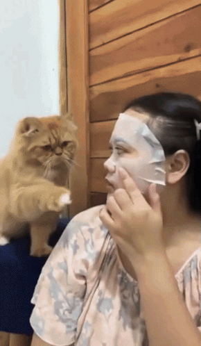 A Woman Is Putting A Face Mask On Her Face While A Cat Looks On GIF