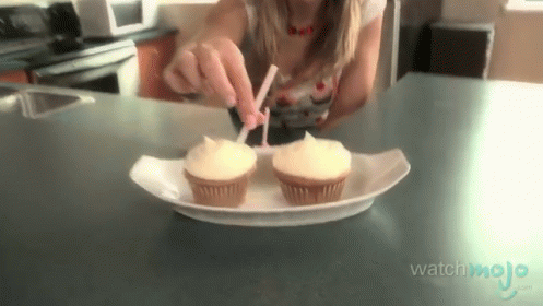 A Woman Is Putting Frosting On Two Cupcakes That Are On A Paper Plate GIF