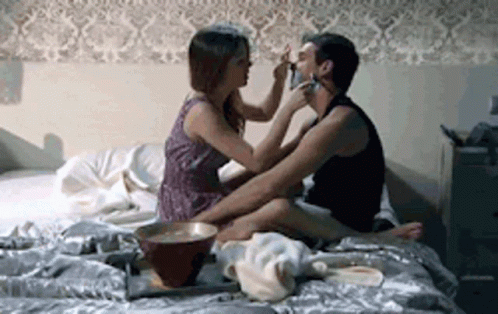 A Woman Is Putting Makeup On A Man 'S Face While They Are Sitting On A Bed . GIF