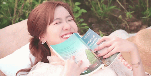 A Woman Is Reading A Book And Smiling While Laying On A Blanket GIF