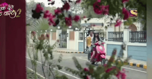 A Woman Is Riding A Scooter Down A Street With Flowers In The Foreground And A Sony Channel Logo In The Background GIF