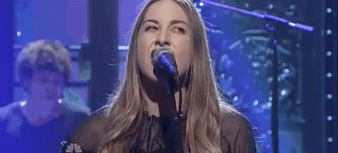 A Woman Is Singing Into A Microphone On A Stage With A Man Playing A Piano In The Background . GIF