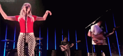 A Woman Is Singing Into A Microphone While A Man Plays A Guitar On A Stage . GIF