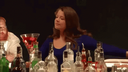 A Woman Is Sitting At A Bar Surrounded By Bottles Of Liquor . GIF
