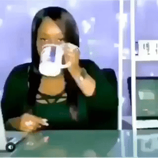 A Woman Is Sitting At A Desk Drinking From A Mug GIF