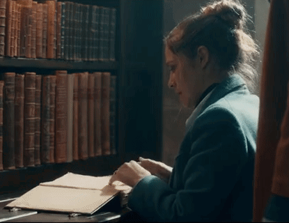 A Woman Is Sitting At A Desk In Front Of A Bookshelf In A Library Looking At A Book . GIF