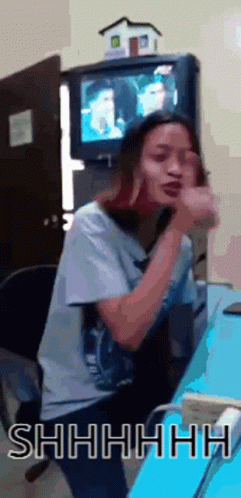 A Woman Is Sitting At A Desk In Front Of A Television And Making A Shhh Gesture GIF
