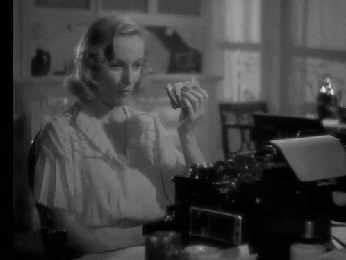 A Woman Is Sitting At A Desk In Front Of A Typewriter And Holding Something In Her Hand GIF