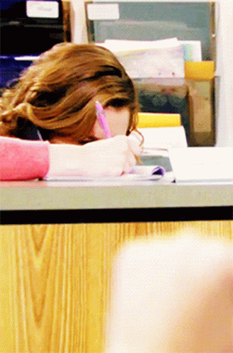 A Woman Is Sitting At A Desk With Her Head Down Writing In A Notebook GIF