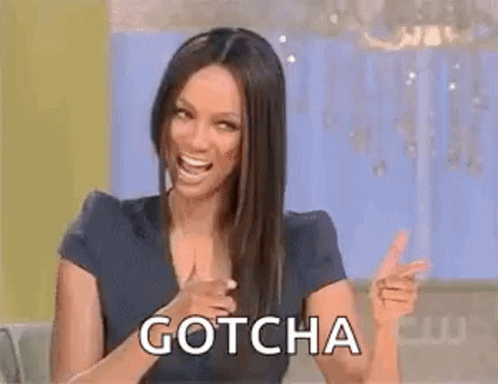 A Woman Is Sitting At A Table And Pointing At The Camera While Laughing And Saying `` Gotcha '' . GIF