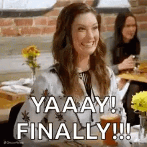 A Woman Is Sitting At A Table Drinking A Beer And Smiling While Another Woman Looks On . GIF