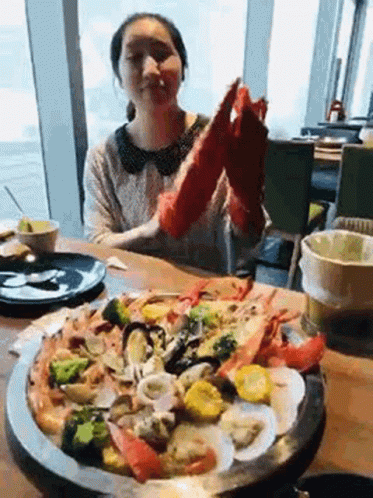A Woman Is Sitting At A Table Eating A Pizza With Seafood And Vegetables . GIF