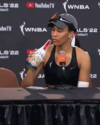 A Woman Is Sitting At A Table Holding A Bottle Of Ultra Light Beer GIF