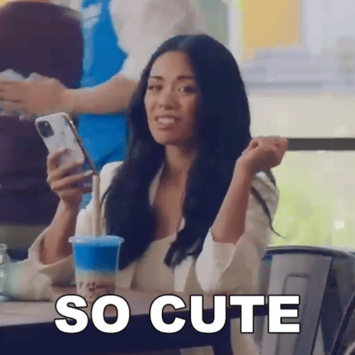 A Woman Is Sitting At A Table Holding A Cell Phone And A Cup Of Blue Liquid . GIF