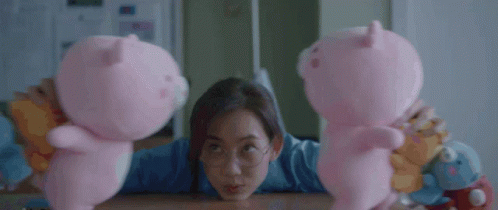 A Woman Is Sitting At A Table Holding Two Pink Pig Stuffed Animals GIF