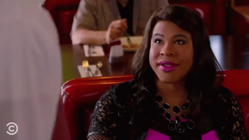 A Woman Is Sitting At A Table In A Diner Wearing A Pink Top And Black Lace Top . GIF