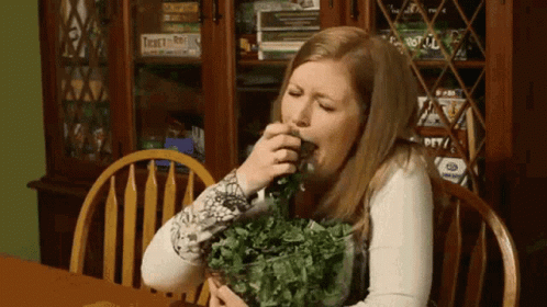 A Woman Is Sitting At A Table With A Bowl Of Greens In Front Of A Bookcase With Twenty-One On It GIF