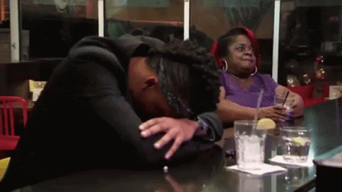 A Woman Is Sitting At A Table With A Drink In Her Hand And A Man Is Laying On The Table With His Head Down . GIF